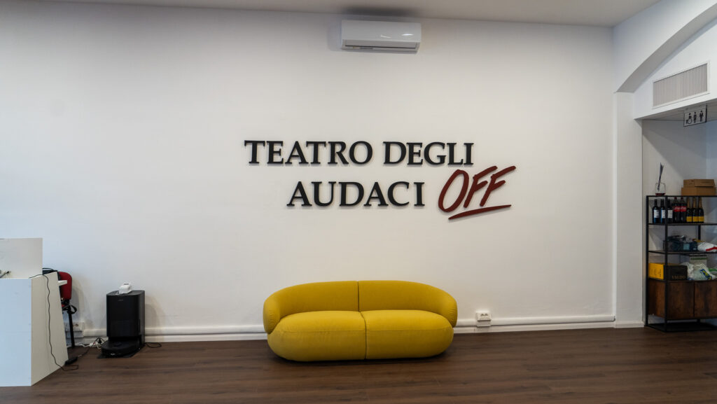 Il foyer del Teatro degli Audaci Off con l’insegna a parete e il divano giallo